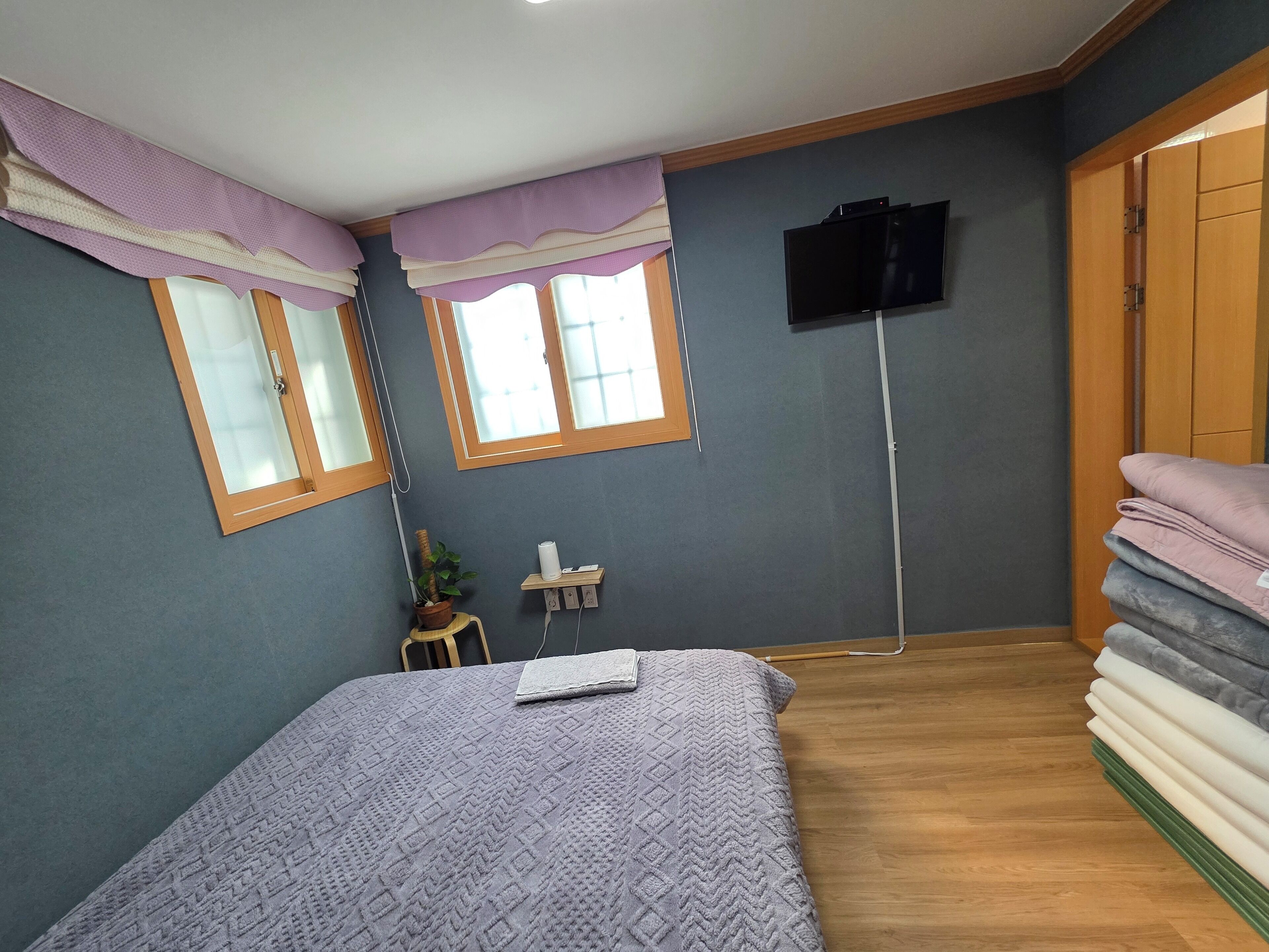 Ein Schlafzimmer mit einem Bett, zwei mit Vorhängen versehenen Fenstern, einem an der Wand befestigten Fernseher, einem kleinen Tisch mit einer Pflanze und einem Stapel gefalteter Decken.