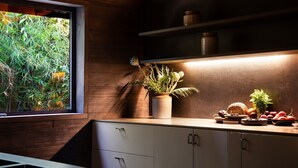 A modern kitchen with wooden cabinetry, a window with a view of greenery, and a countertop with a potted plant and food items.