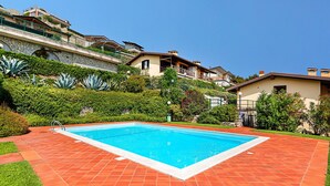 A rectangular swimming pool with a tiled deck, surrounded by lush greenery and residential buildings in the background.
