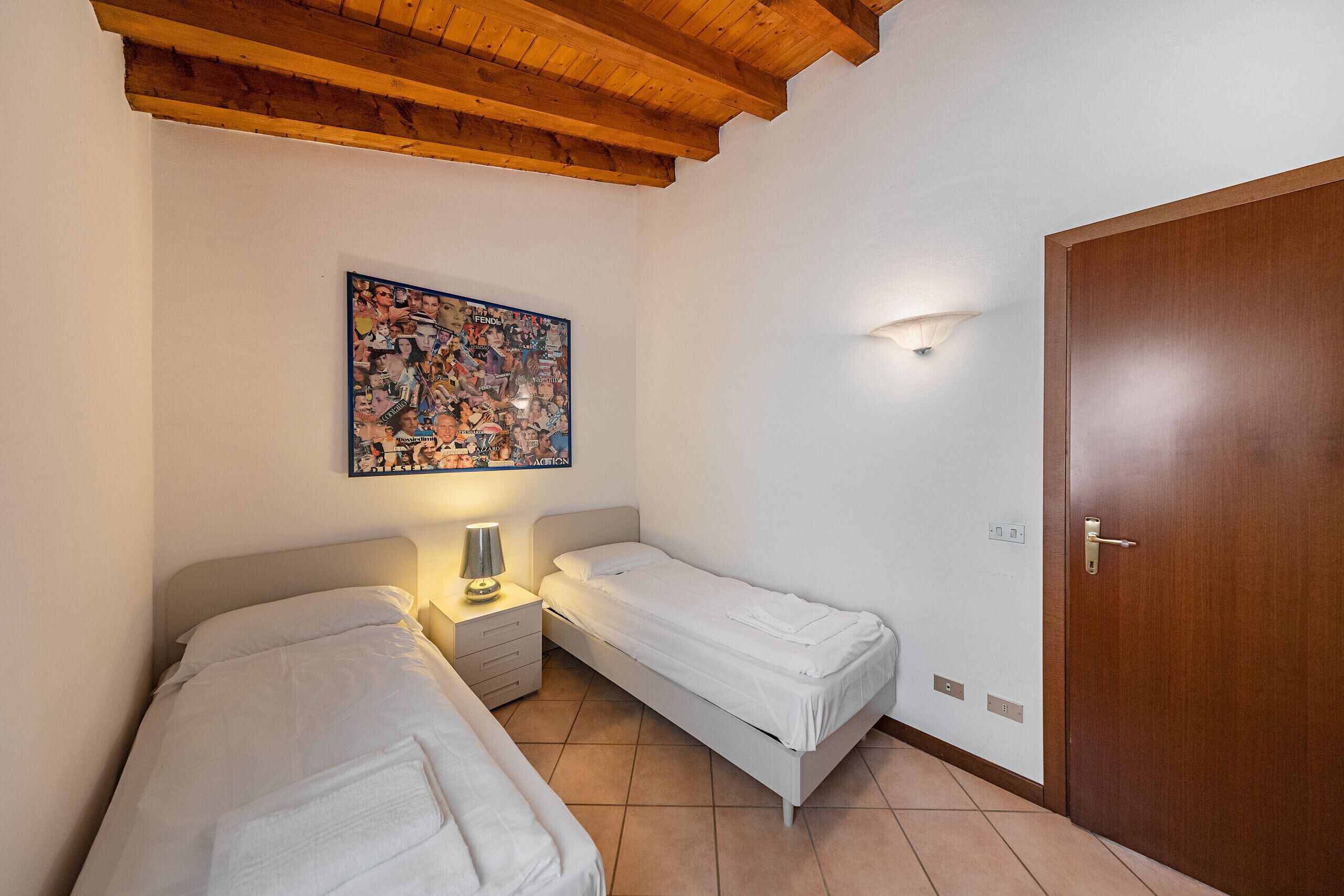 A bedroom with two single beds, a wooden ceiling, a wall-mounted light, and a door.