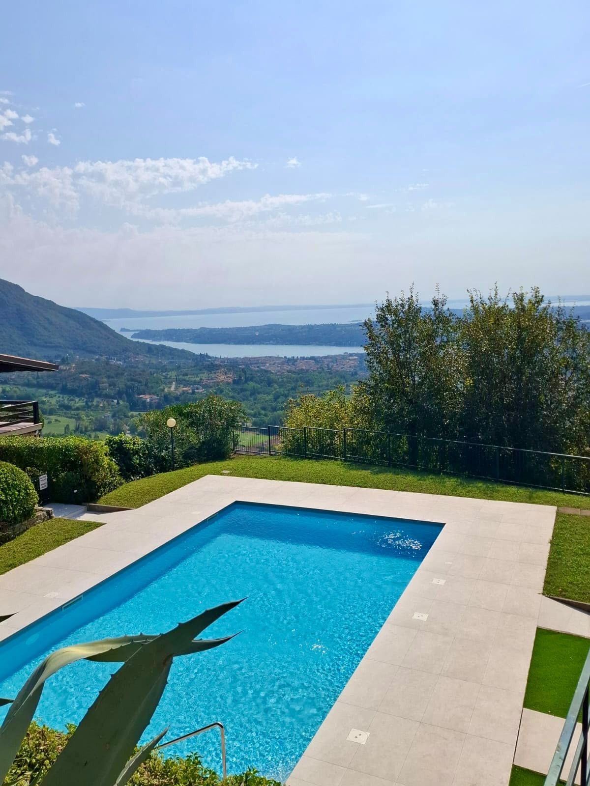 A rectangular swimming pool with clear blue water, surrounded by a paved deck. In the background, there is a view of a lake and distant mountains under a clear sky.