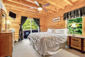 A wooden cabin bedroom with a bed, a ceiling fan, a TV, a window with curtains, and a view of greenery outside.