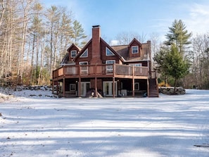 A large, two-story wooden cabin with a balcony, surrounded by a snowy landscape and trees.
