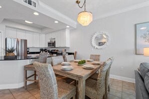 A modern kitchen with white cabinets, a stainless steel refrigerator, and a dining area with wicker chairs and a wooden table. - Updated Luxury Beachfront Condo w/ Pool, Sauna, & Fitness Center | Sleeps 8 (Gulf Shores)