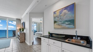 A kitchen with a black countertop, white cabinets, and a large painting of a beach scene. - Updated Luxury Beachfront Condo w/ Pool, Sauna, & Fitness Center | Sleeps 8 (Gulf Shores)