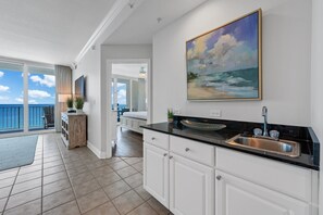 A kitchen with a black countertop, white cabinets, and a large painting of a beach scene. - Updated Luxury Beachfront Condo w/ Pool, Sauna, & Fitness Center | Sleeps 8 (Gulf Shores)