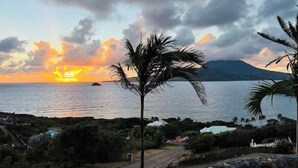 A coastal view with a sunset, palm trees, and a mountain in the background.