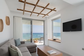 A modern living room with a sofa, a coffee table, and a TV. There are large windows offering a view of the ocean.