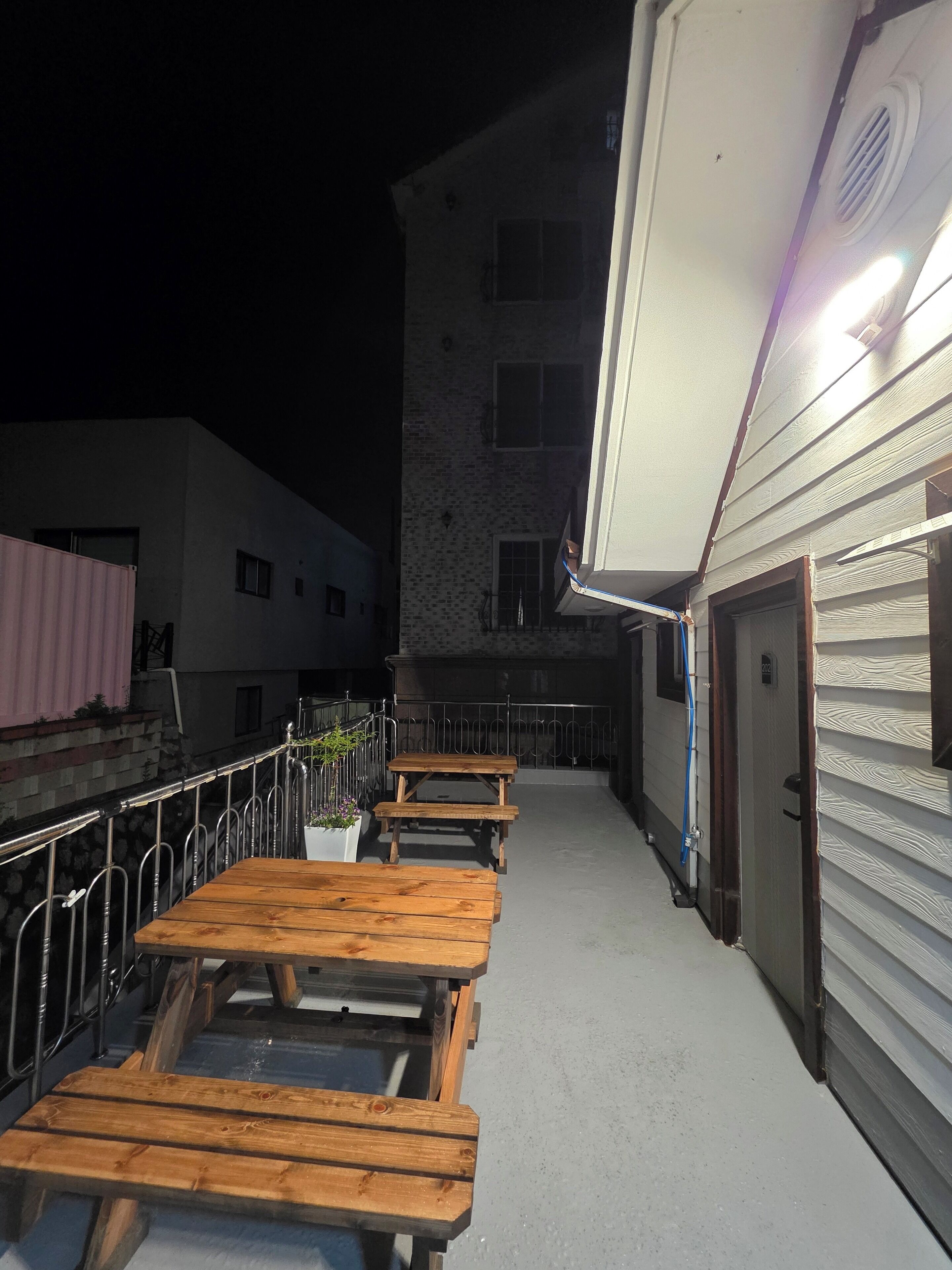 An outdoor patio area with wooden tables and chairs, a metal railing, and a building in the background.