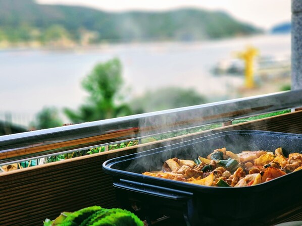 A view of a lakeside balcony with a grill cooking food. - Beach Getaway (Suite 201) for 1 - 3 guests: Perfect Location & Great Views. (Geoje-si)