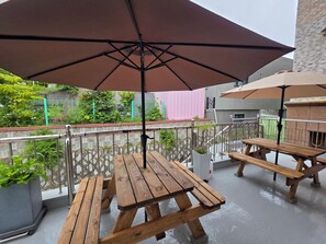 A patio area with wooden tables and benches, a large umbrella, and a view of a garden and buildings in the background. - Beach Getaway (Suite 201) for 1 - 3 guests: Perfect Location & Great Views. (Geoje-si)