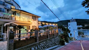 A two-story building with a balcony, outdoor seating, and a view of the sea. - Beach Getaway (Suite 201) for 1 - 3 guests: Perfect Location & Great Views. (Geoje-si)