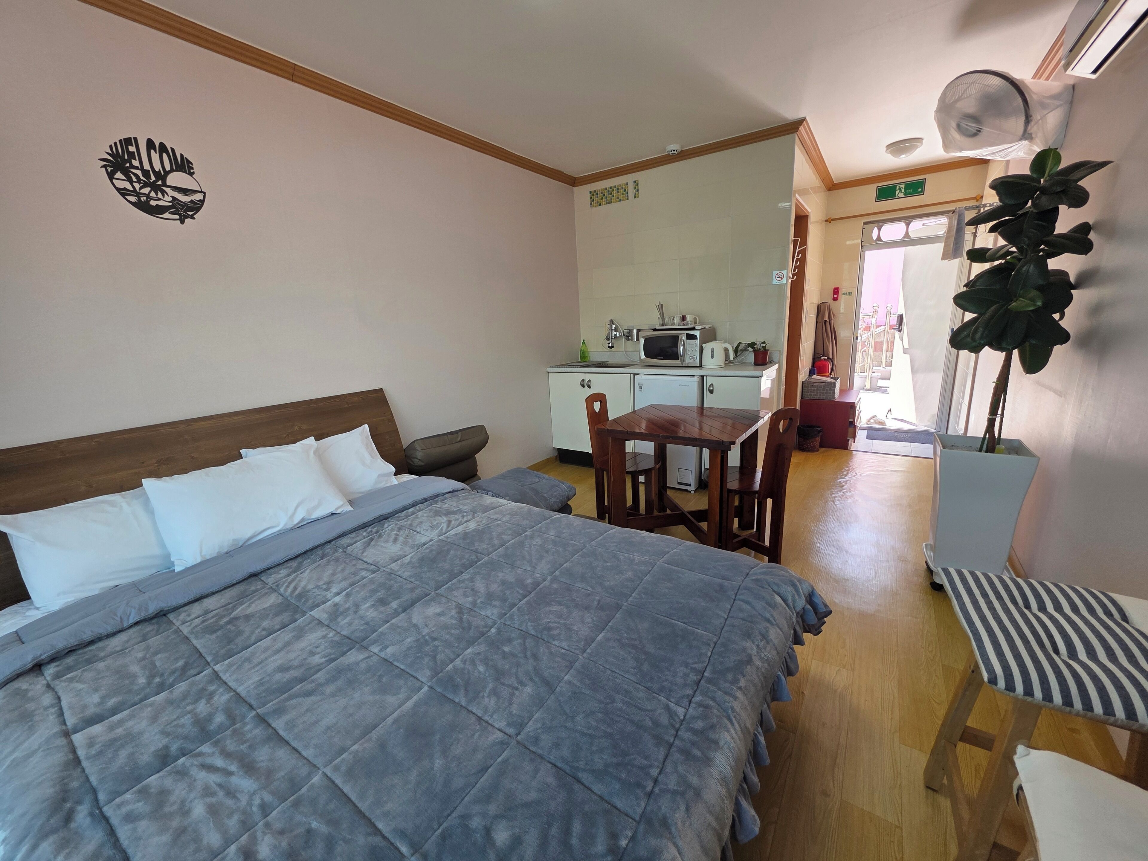 A bedroom with a bed, a small table, and a chair. There is a kitchenette in the background with a microwave and a sink.