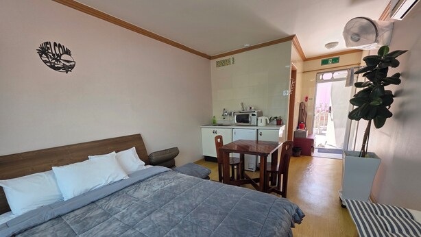Quarto com uma cama, uma mesinha e uma cadeira. É possível ver uma cozinha compacta ao fundo, com um micro-ondas e uma pia.