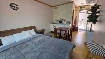 A bedroom with a bed, a small table, and a chair. There is a kitchenette in the background with a microwave and a sink.