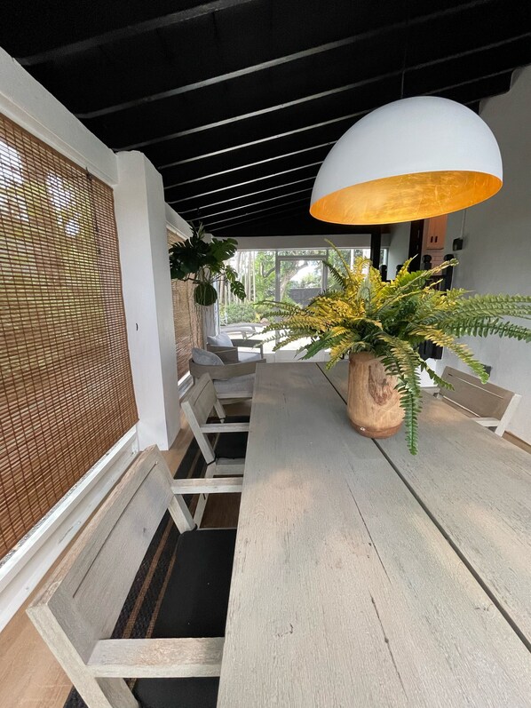 A dining area with a wooden table, white chairs, a large potted fern, and a hanging light fixture.