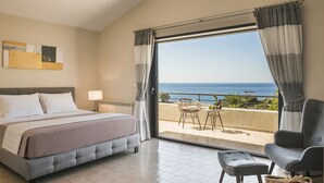 Un dormitorio con cama, mesita de noche, lámpara, vista al mar a través de un ventanal grande, y un balcón con una silla y una mesa.