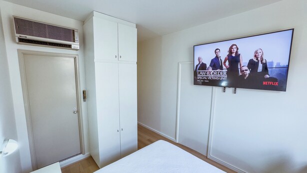 A hotel room with a bed, a wardrobe, a television displaying a show, and a wall-mounted air conditioner. - City Studio | Netflix Premium 4K | 65" LG QLED TV | Queen Memory Foam Bed (Adelaide)