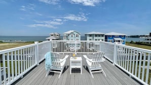 Eine Holzterrasse mit weißen Geländern, zwei weiße Adirondack-Stühle und ein Tisch mit einem Getränk. Im Hintergrund sind mehrere Strandhäuser mit blau-weißen Fassaden zu sehen.