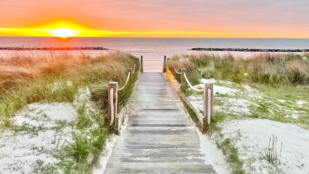 Ein hölzerner Steg führt zu einem Strand, im Hintergrund ist ein Sonnenuntergang zu sehen.