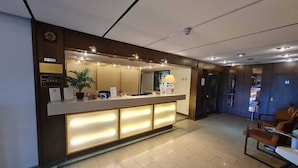 A hotel reception area with a reception desk, a plant, a lamp, and a seating area with chairs.