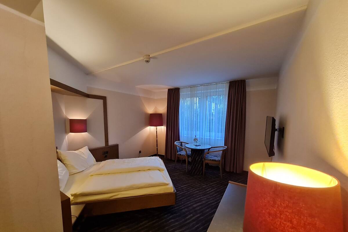 A hotel room with a bed, bedside lamps, a desk with a chair, a window with curtains, and a wall-mounted TV.
