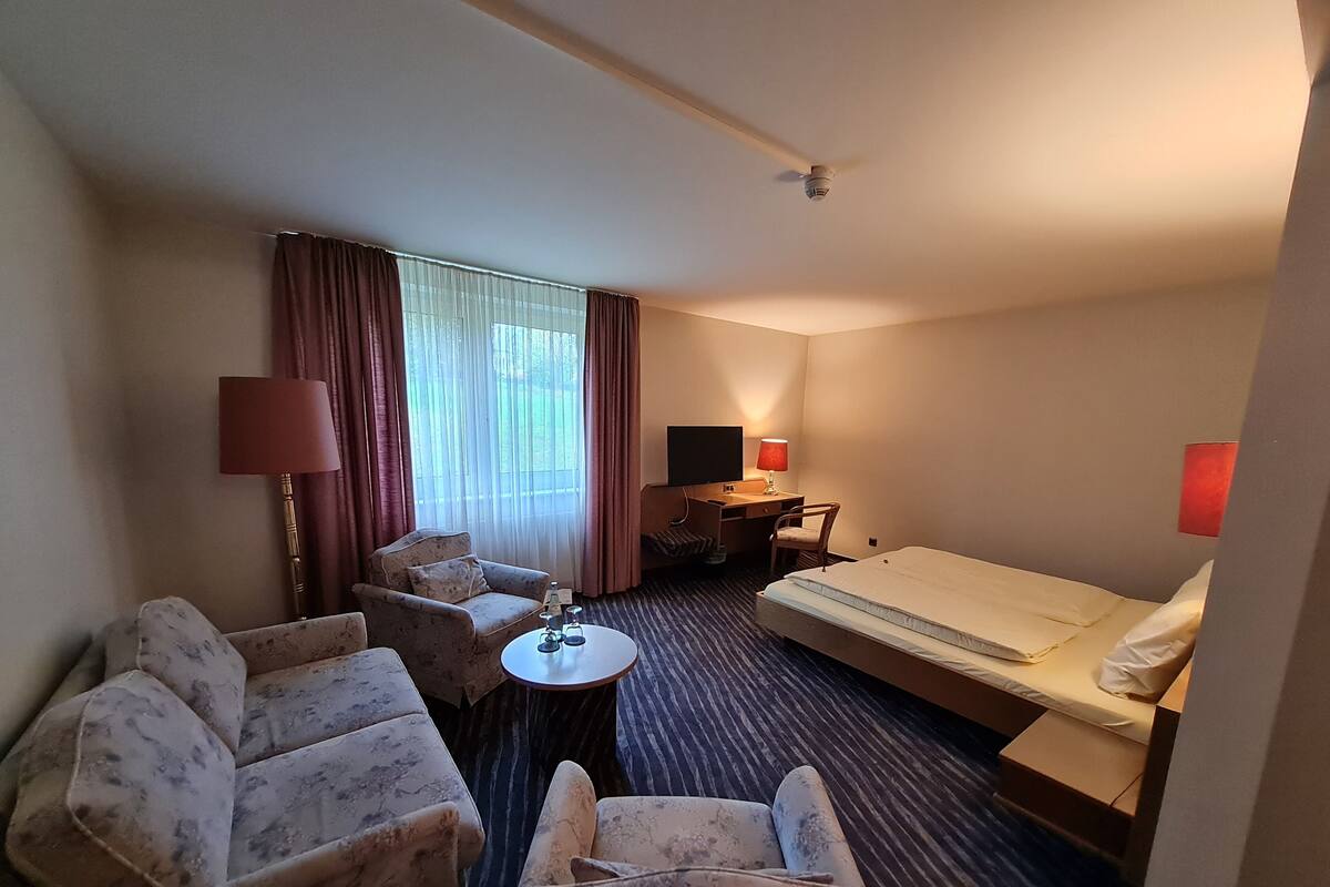 A hotel room with a bed, two armchairs, a small table, a desk with a computer, and a window with curtains.