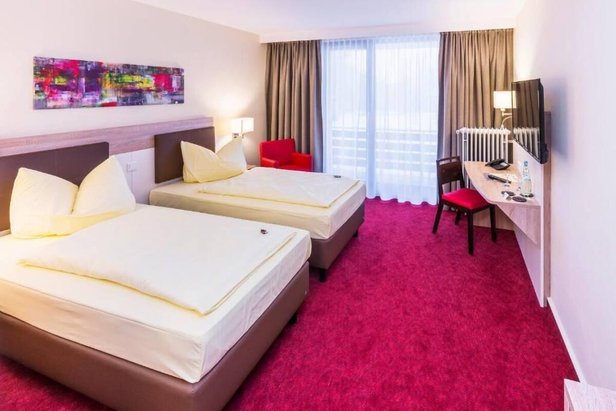 A hotel room with two beds, a desk, a chair, a television, and a window with curtains.