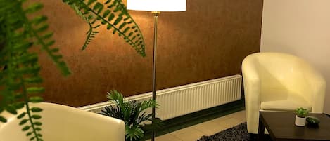 Una habitación con un sillón blanco, una lámpara de pie, una pequeña mesa con una planta y una grande planta de helecho.