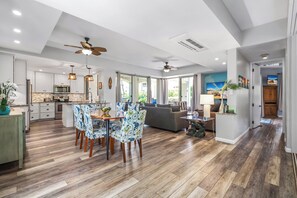 A spacious living area with a dining table, chairs, and a sofa. There is a kitchen in the background with cabinets and a microwave.