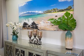 Coastal-inspired decor features a large beach scene painting, a vase with white flowers, and a geometric-patterned console table.