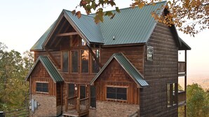 A log cabin with a stone foundation, a green metal roof, and a wooden deck. - Gatlinburg Luxury Cabin, 4 Master Bdrms, Theater, Game Rm, Hot Tub, Perfect! (Sevierville)