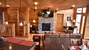 A cozy cabin interior with a stone fireplace, wooden furniture, a dining area, and a staircase. - Gatlinburg Luxury Cabin, 4 Master Bdrms, Theater, Game Rm, Hot Tub, Perfect! (Sevierville)