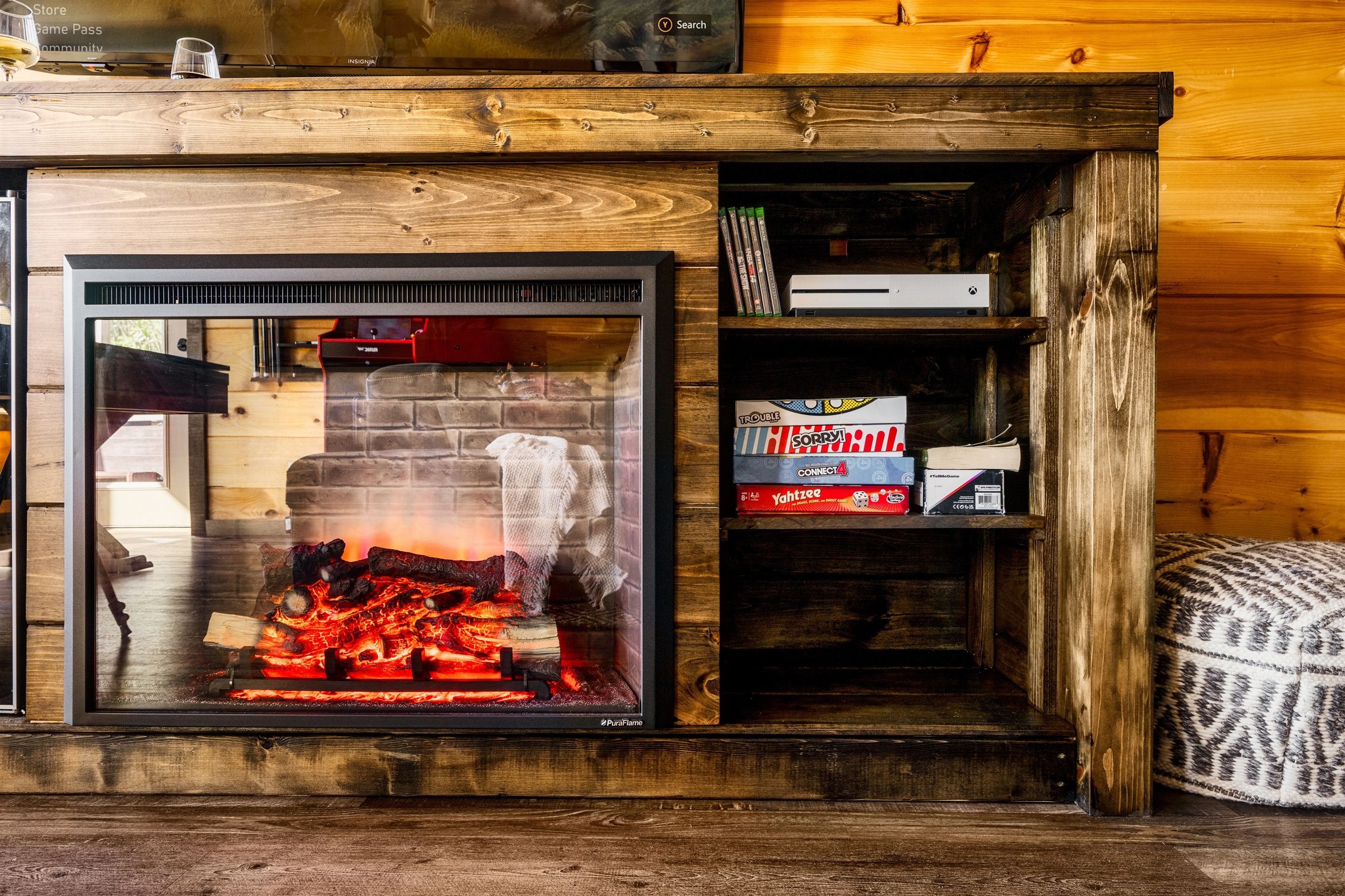 A fireplace with a glass front, a shelf with books and a box labeled "Trouble," and a patterned ottoman.