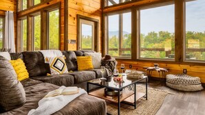 Ein gemütliches Wohnzimmer mit einer Ledersofa, einem Couchtisch und großen Fenstern mit Blick auf Grünflächen und Berge.