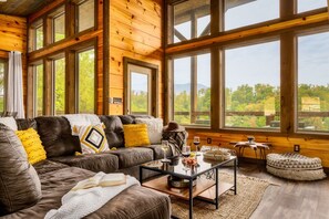 A cozy living room with a leather sofa, a coffee table, and large windows offering a view of greenery and mountains.