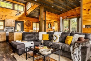 A cozy cabin interior with a living area featuring a leather sofa, a coffee table, and a kitchen area with wooden cabinets and a countertop.