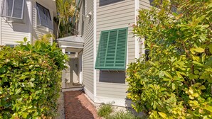 A narrow brick pathway leading to a building with white siding and green shutters, surrounded by lush greenery. - ISLAND OASIS ~ Stylish 2 Bedroom, 2 Bath Home w/ Private Heated Pool! (Key West)