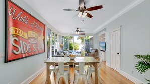 A dining area with a wooden table and white chairs, a ceiling fan, and a large poster on the wall. - ISLAND OASIS ~ Stylish 2 Bedroom, 2 Bath Home w/ Private Heated Pool! (Key West)