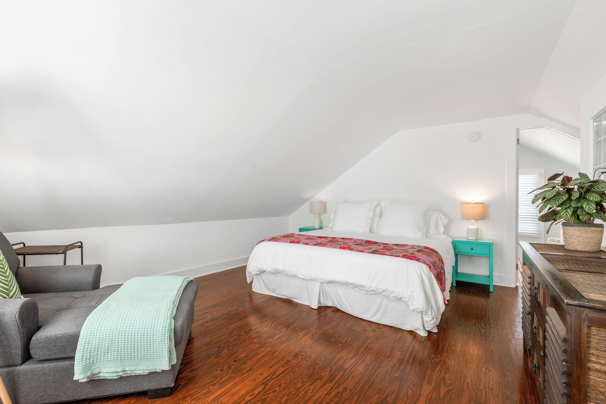 A bedroom with a bed, a chair, a nightstand, a dresser, and a potted plant.