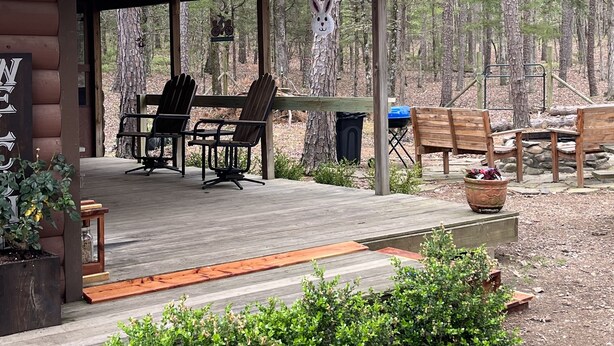 A wooden cabin with a covered porch, outdoor seating, and a forest backdrop. - Fenced for your fur babies! 2 cabins on property for 1 price! (Mena)