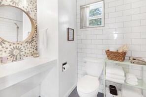 A bathroom with a white toilet, a glass shelf with towels, a wicker basket, and a framed picture on the wall.
