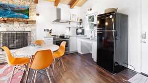 Una cucina moderna con parete in pietra, travi in legno, piano cottura integrato, frigorifero nero e sedie arancioni attorno a un tavolo bianco.