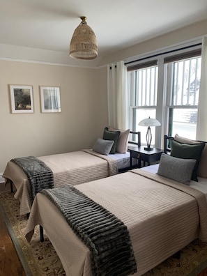 A hotel room with two beds, a large window with curtains, a hanging light fixture, and two framed pictures on the wall.