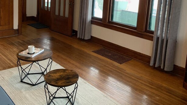 A spacious living room with wooden floors, a large window with curtains, a wooden door, and a hanging light fixture.