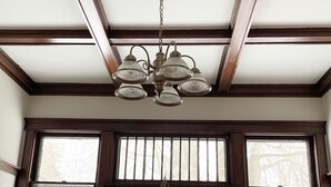 A dining room with a wooden table, four chairs, a chandelier, and large windows with a view of trees.