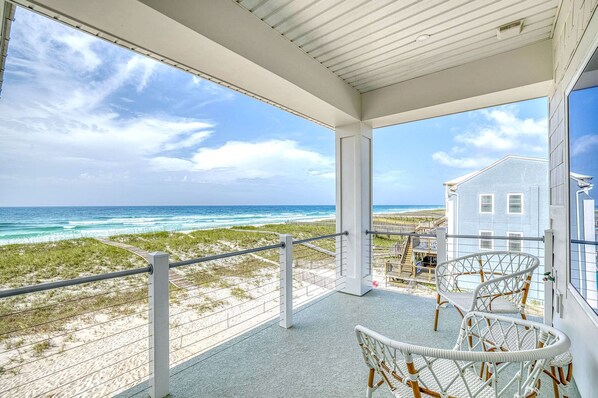 A balcony with a view of the ocean and beach, white wicker chairs, and a building in the background.