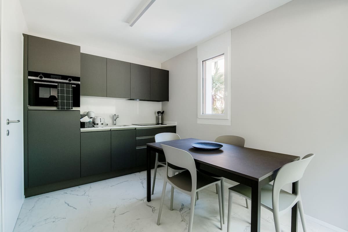 Une cuisine moderne avec des meubles de couleur sombre, un plan de travail en marbre et un coin repas comprenant une table sombre et des chaises blanches.