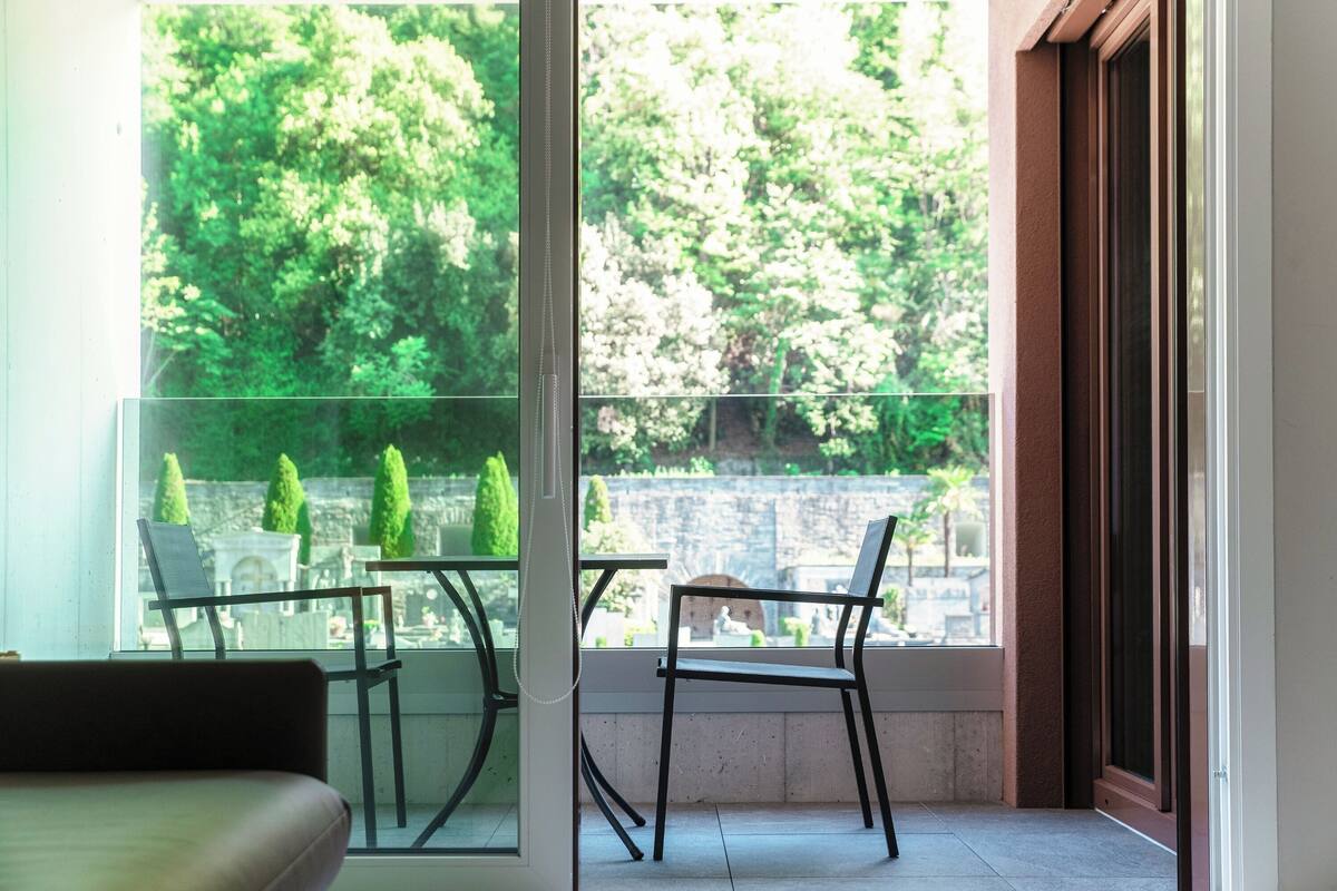 Un balcon avec une porte vitrée, deux chaises et une vue sur la verdure et un mur de pierre.