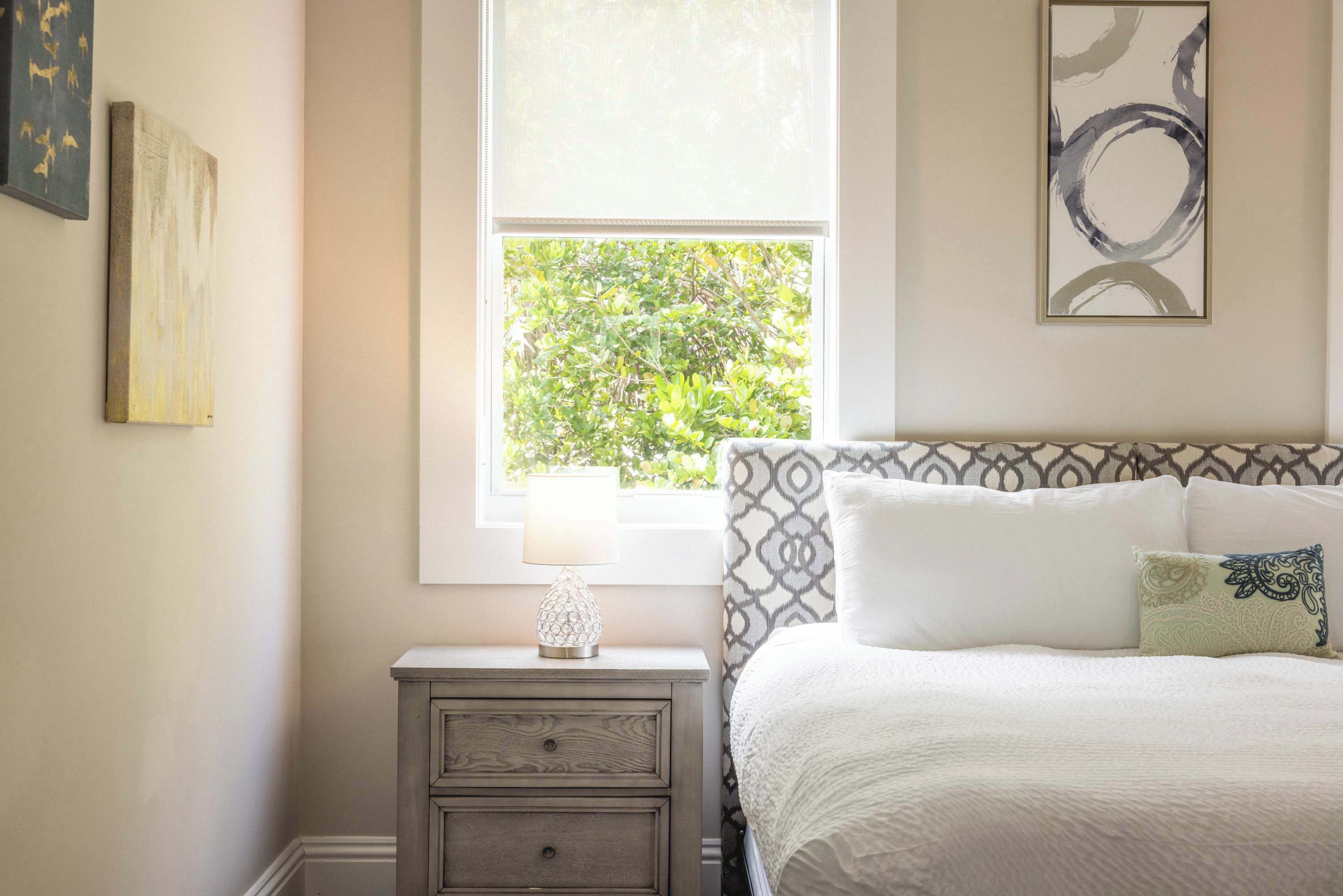 A bedroom with a bed, a nightstand, a window with a view of greenery, and a wall with abstract art.